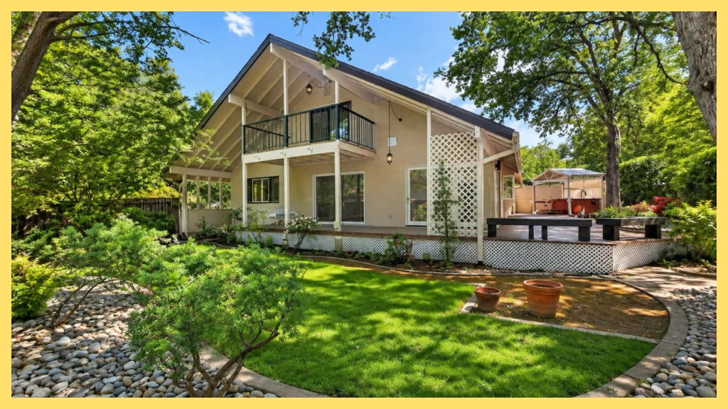 A spacious custom home in Wexford Estates, Granite Bay, California, featuring vaulted rooflines, a covered patio, upper level balcony, expansive backyard deck, and mature oak trees. The landscaped yard offers lush green lawn, garden beds, and outdoor entertaining space, reflecting the peaceful, established setting of Wexford Estates, a desirable neighborhood known for large lots, privacy, and family friendly living.